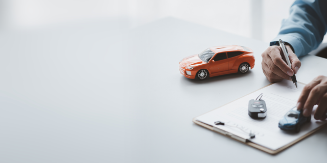 A person at the corner of a desk is signing vehicles papers. We see only his arms and hands, he is holding a pen. Two miniature cars are on the desk.