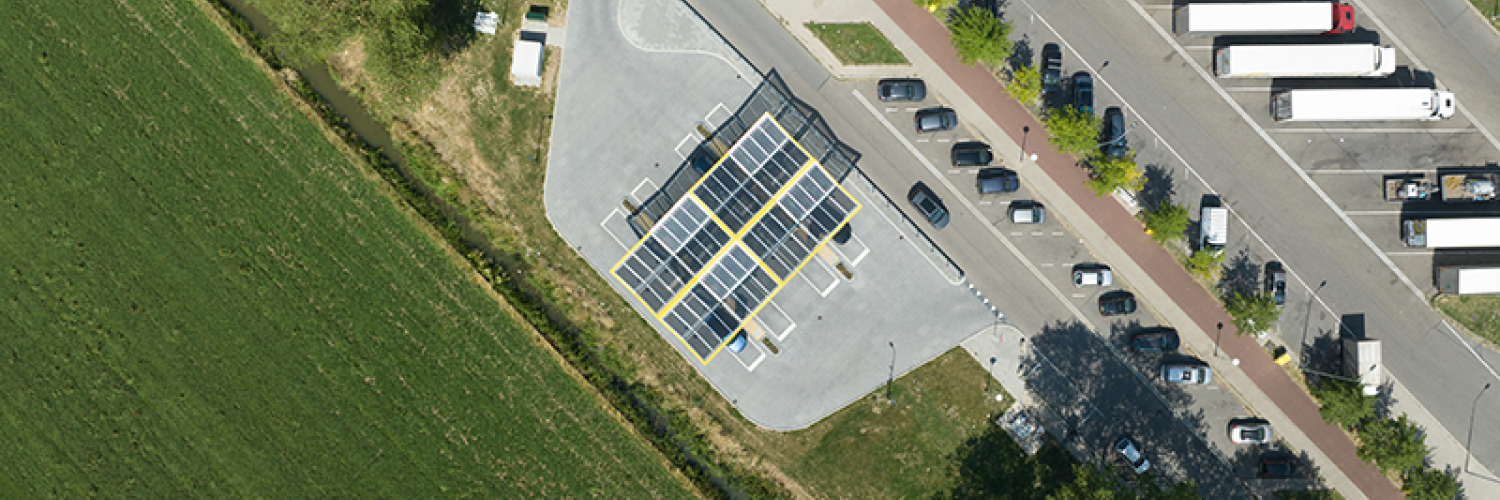 A view from the sky of an EV charging station in a parking lot. The station is covered by a roof composed of solar panels