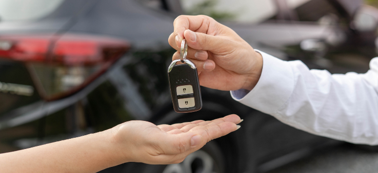 Two persons are in front of a car. There is a close up of their hands, the person on the right is giving car keys to the person on the left