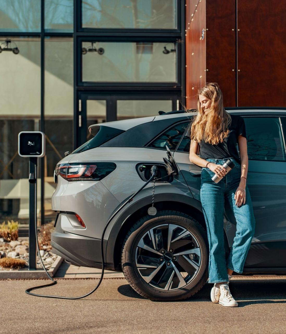 A person leaning against her car that is currently charging. The person is looking at the charging port and smiling