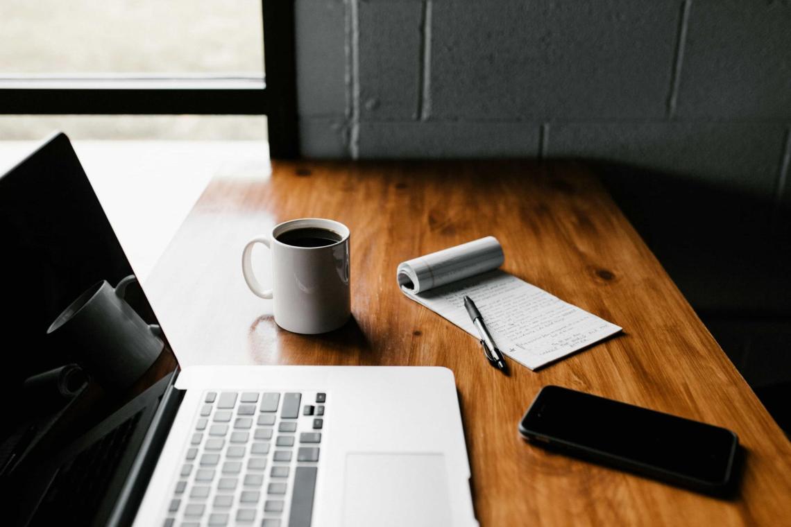 Table with computer a laptop, a smartphone, a mug with coffee and a notebook with a pen are on a wooden desk
