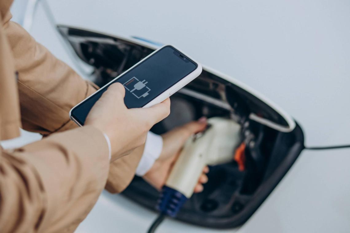 EV charging up close with a view of a phone on the first layer of the picture, we can see a person holding a phone with its right hand. The screen shows a battery loading. With its left hand, the person is holding a charging cable for an electric car