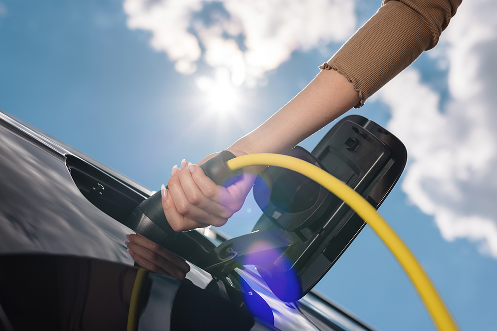 Person plugging an electrified vehicle A close up from below of the hand of a person holding a charger and plugging it in the vehicle. In the background, you can see the sun and the blue sky with a cloud