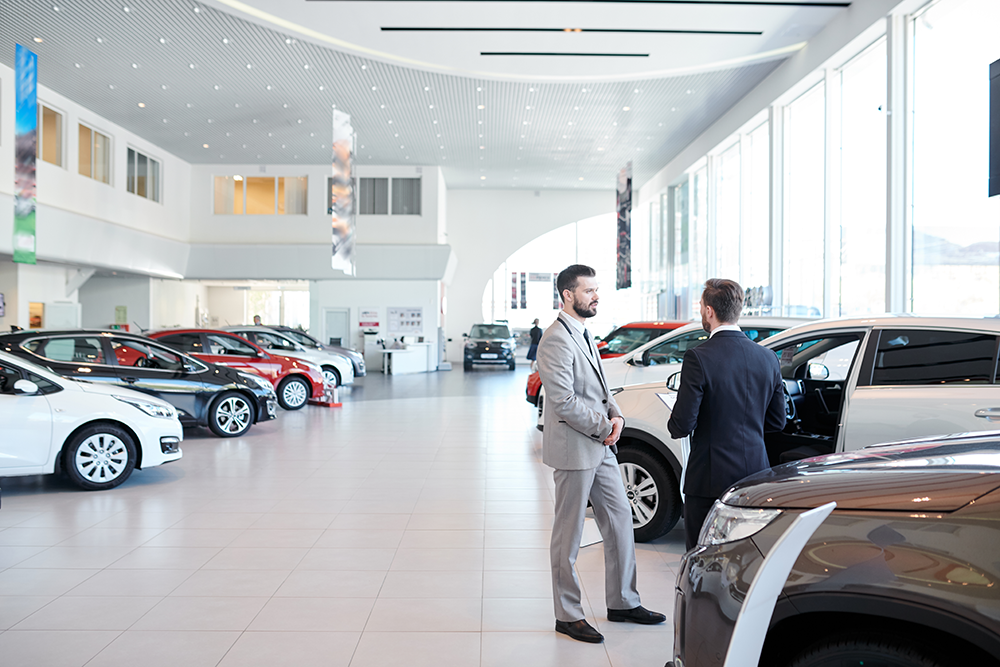 A view from inside a dealership with multiple vehicles parked. Two professionals are discussing in front on a car with the front door open in the front part of the picture.